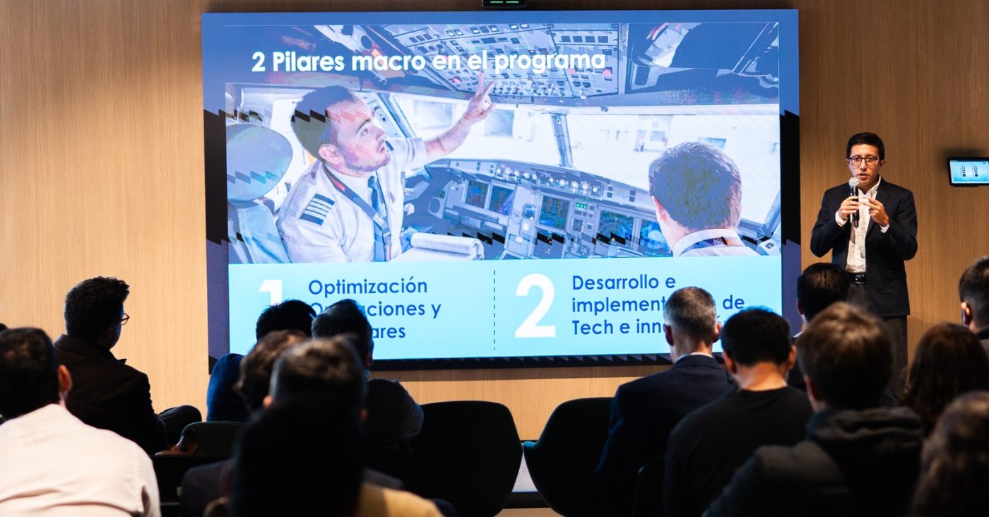 Speaker in a suit presenting to an audience beside a large screen displaying a Spanish slide: '2 Pilares macro en el programa'.