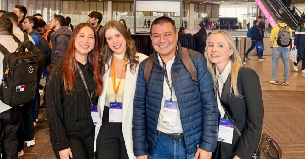 Four people smile for a group photo at a conference, wearing lanyards with badges in a busy indoor venue.