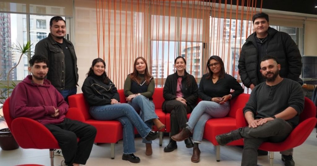 Group of eight people seated on red couches in a bright lounge area, posing for a team photo in an office setting.