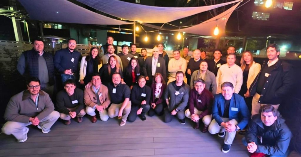Large group of people posing for a group photo on a rooftop terrace with string lights at night, everyone wearing name tags.