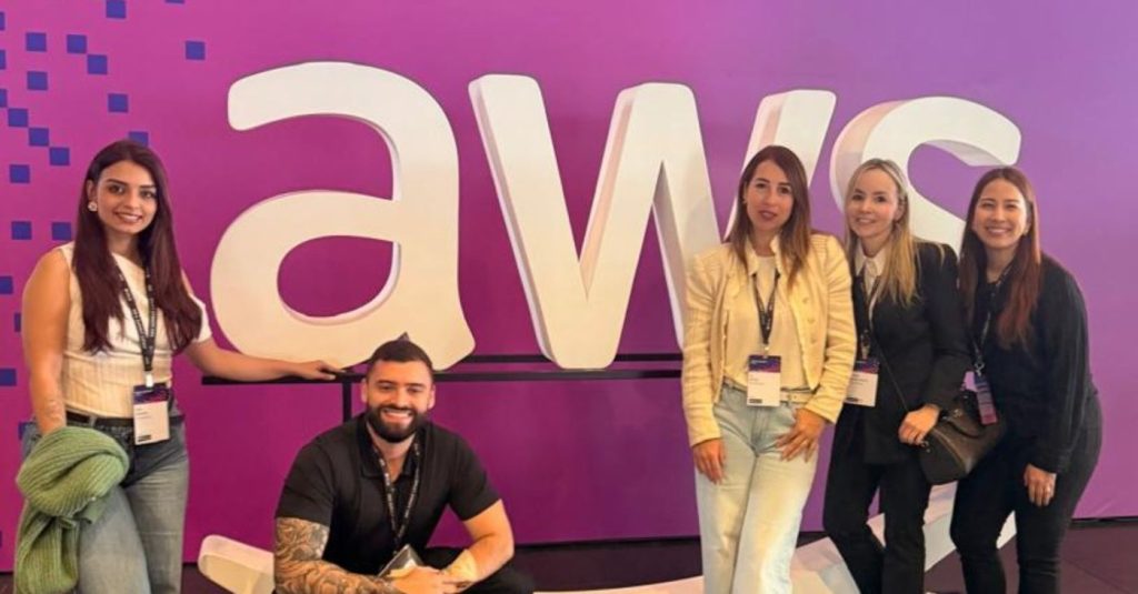 Five people posing for a photo in front of a large white 'aws' logo on a purple wall at a conference; a tattooed man kneels in front, flanked by four women wearing lanyards.