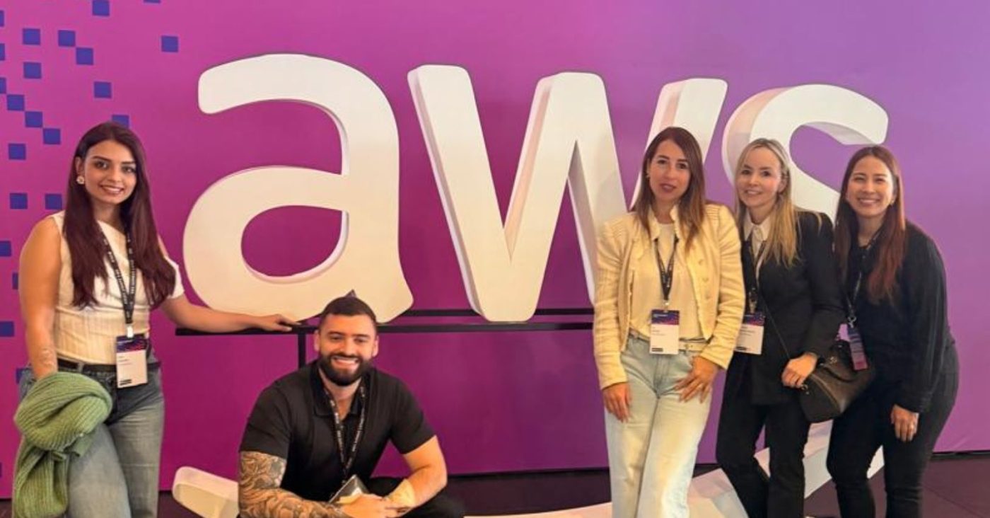 Five people posing for a photo in front of a large white 'aws' logo on a purple wall at a conference; a tattooed man kneels in front, flanked by four women wearing lanyards.
