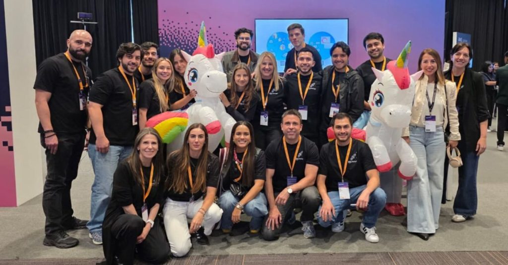 Group of people posing for a photo with two inflatable unicorn mascots at a conference or event hall.